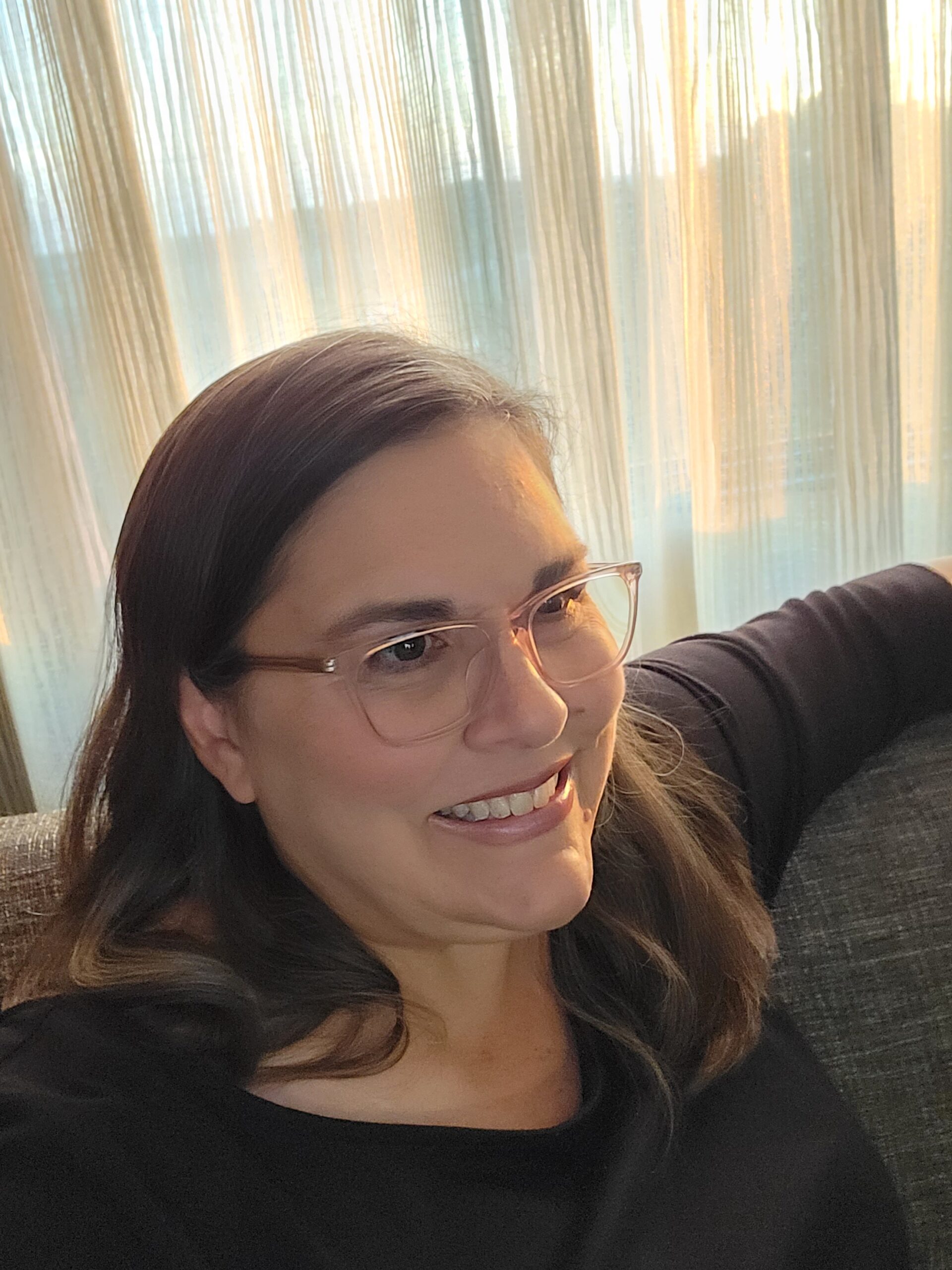 Dana Engelbert casually seated on a dark sofa with a light-diffused background.